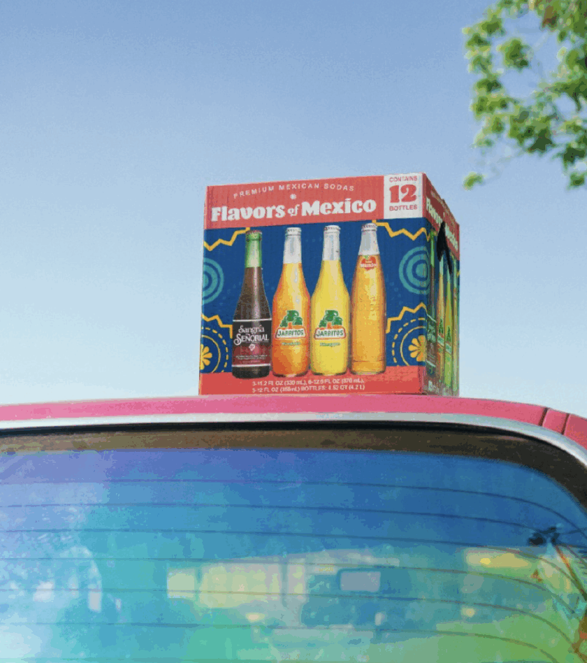 Box of Flavors of Mexico premium sodas, featuring Señorial and Jarritos bottles, sitting on top of a car against a clear blue sky.