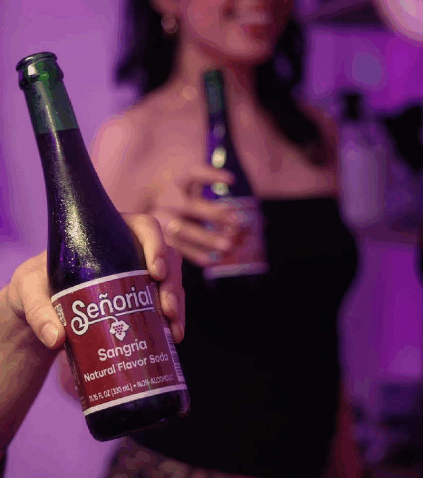 Close-up of a hand holding a chilled Señorial Sangría soda bottle at a party, with a woman in the background also holding a bottle.