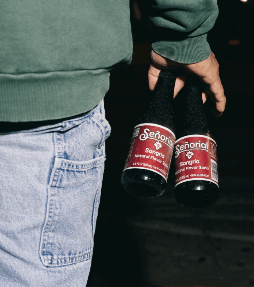 A person holding two chilled bottles of Señorial Sangría soda during a nighttime outing, with denim jeans and a green sweater visible.