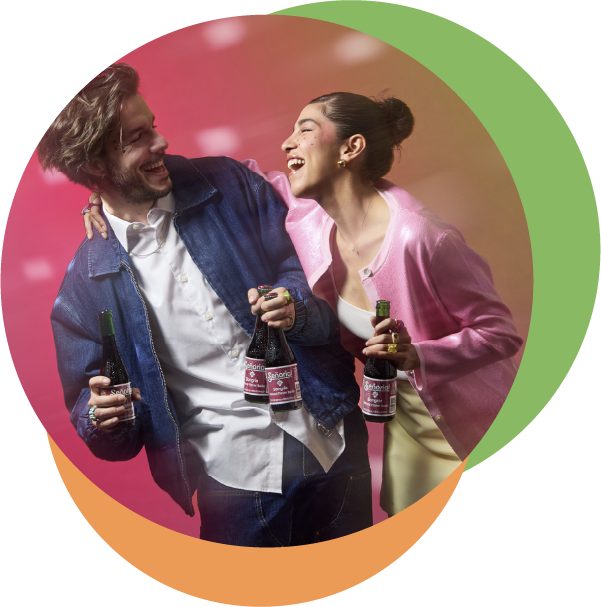 A young man and woman laugh together while holding glass bottles of Señorial Sangría soda against a vibrant pink and orange backdrop.