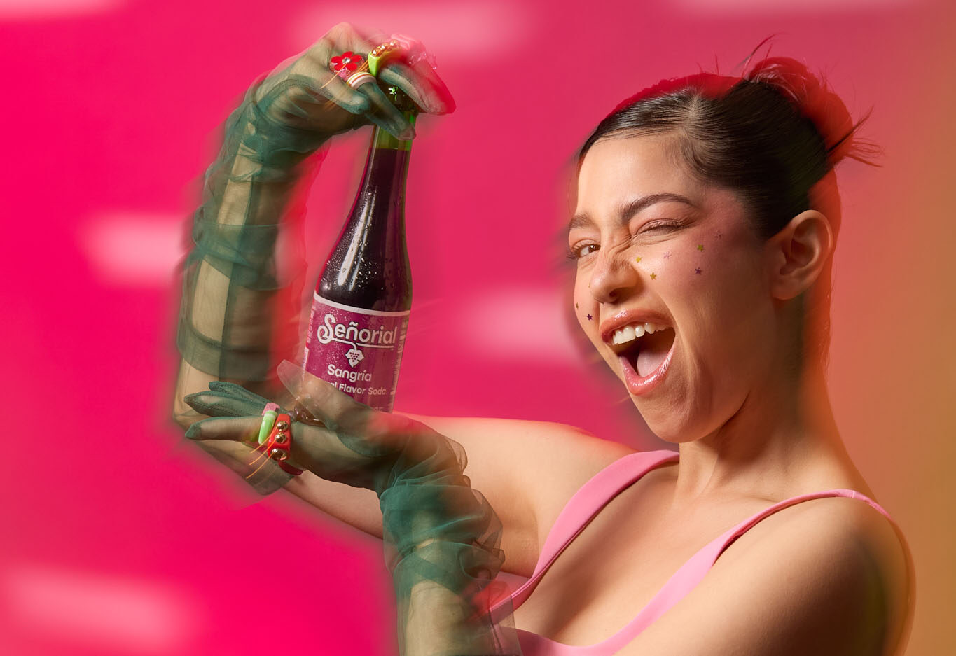 A woman playfully winking while presenting a Sangría Señorial bottle against a vibrant pink background.