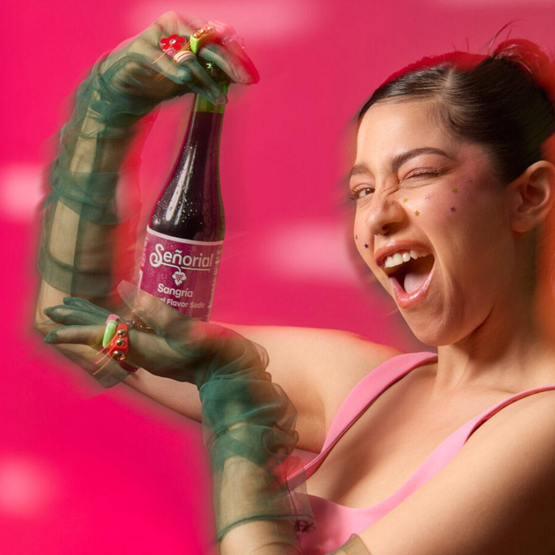 A woman playfully winking while presenting a Sangría Señorial bottle against a vibrant pink background.