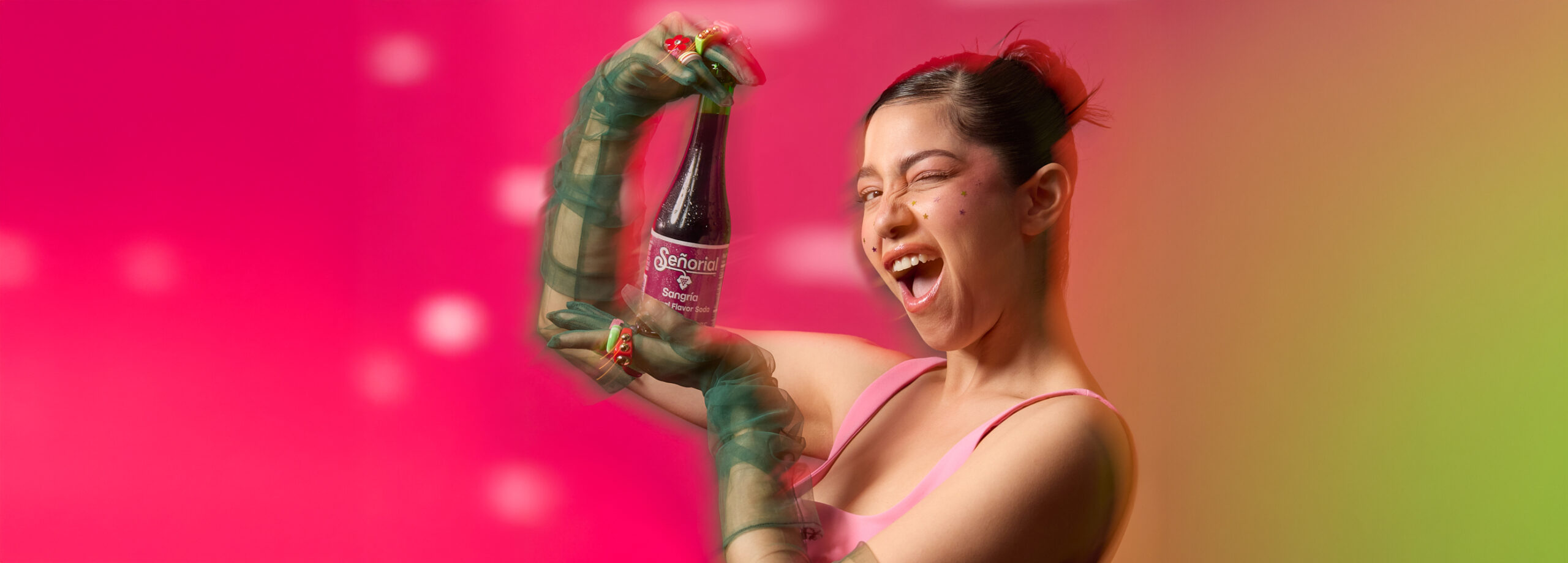 A woman playfully winking while presenting a Sangría Señorial bottle against a vibrant pink background.