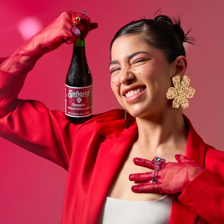 A smiling woman in bold red holding a Sangría Señorial bottle playfully against a vibrant pink-green gradient backdrop.