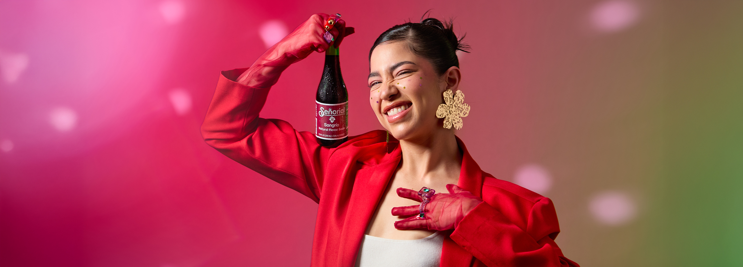 A smiling woman in bold red holding a Sangría Señorial bottle playfully against a vibrant pink-green gradient backdrop.
