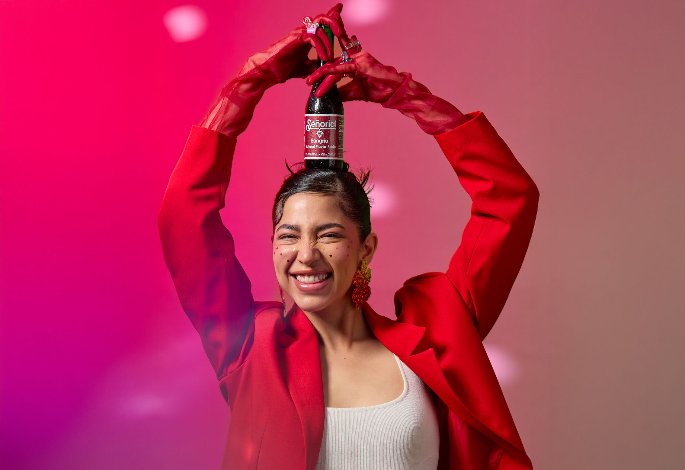 Smiling woman in a red blazer playfully holding a Sangría Señorial bottle above her head against a vibrant pink background.