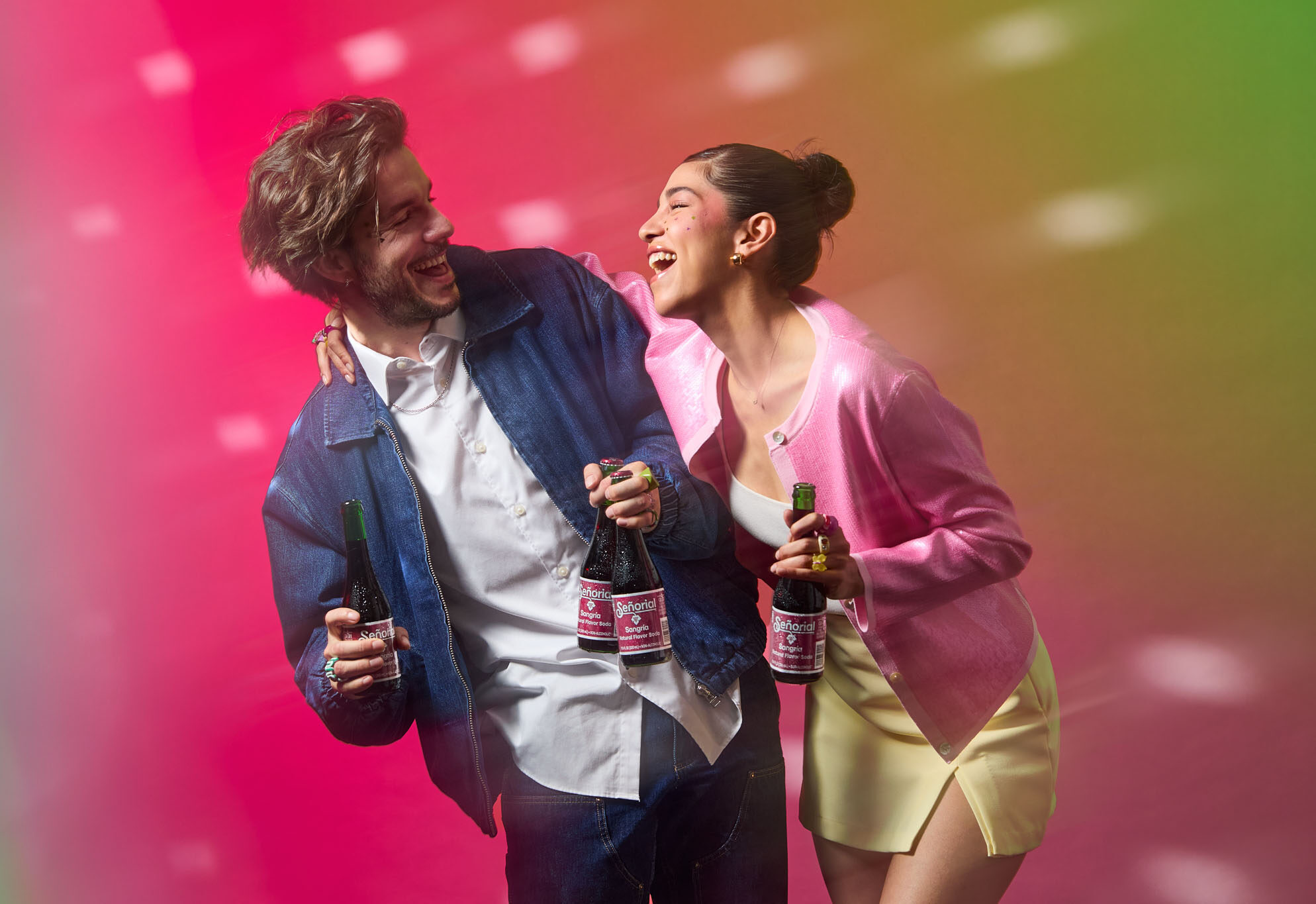 Two friends laughing and holding Sangría Señorial bottles against a vibrant, colorful backdrop full of energy.