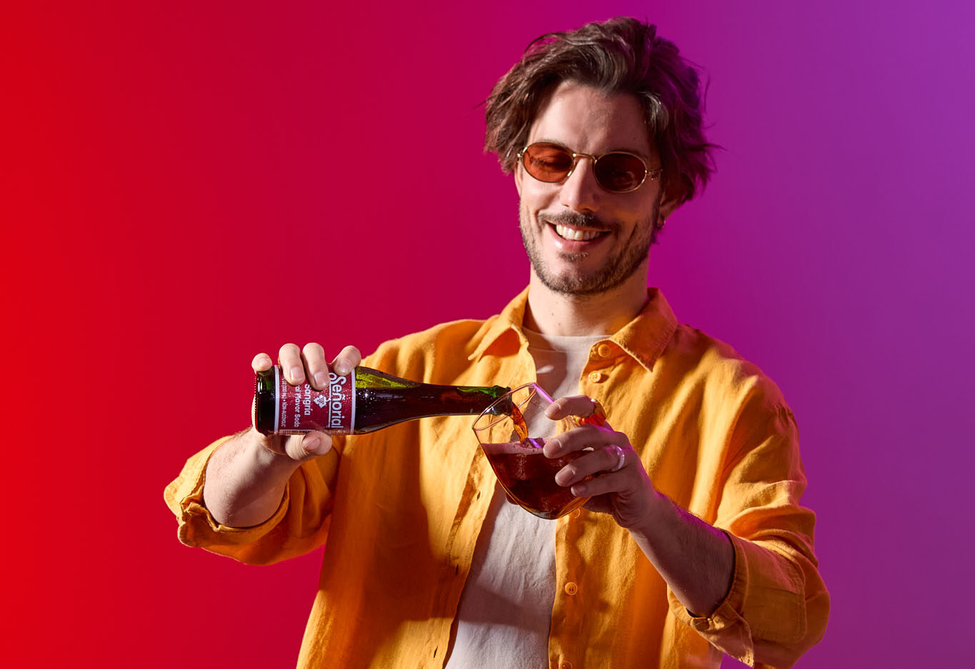 A man smiling as he pours Sangría Señorial into a glass against a vibrant red-pink gradient backdrop.