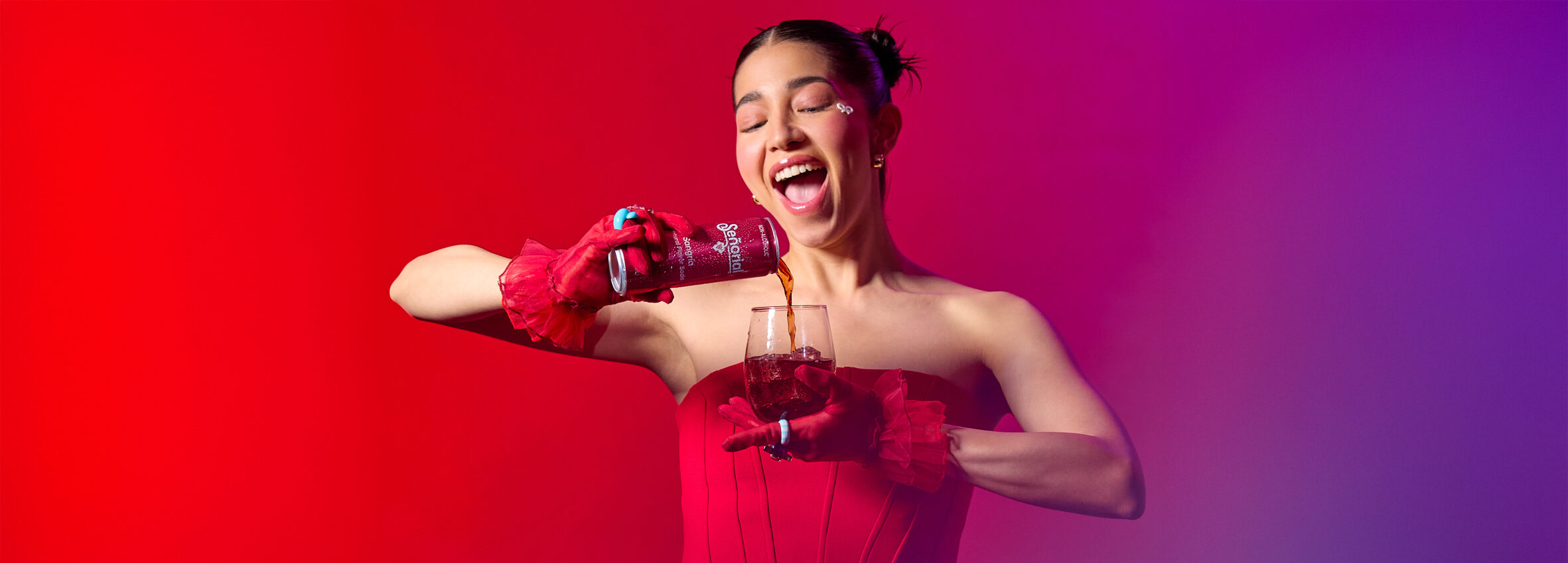 A woman in vibrant red joyfully pouring Sangría Señorial into a glass against a bold red-pink gradient backdrop.