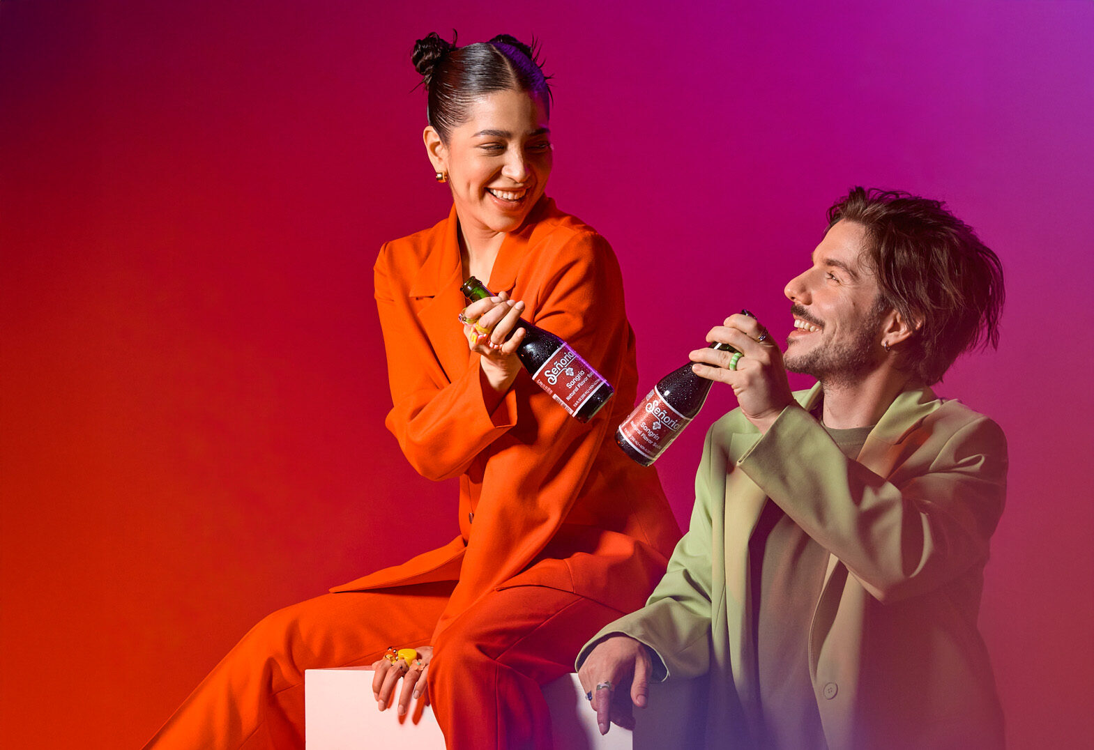 Two people smiling and toasting with Sangría Señorial bottles against a vibrant red-pink gradient backdrop.