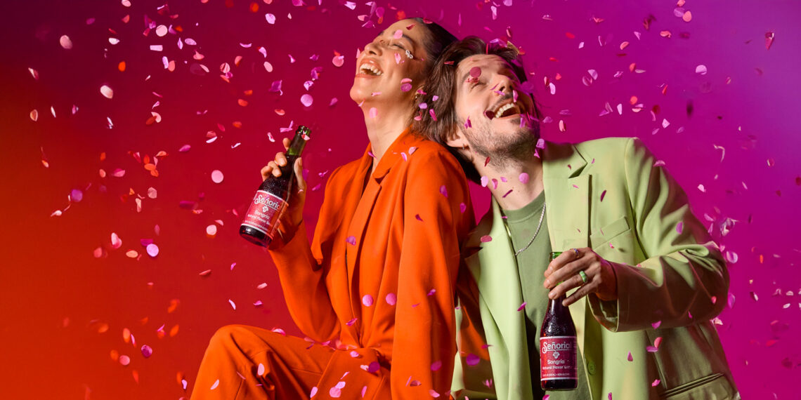 wo people celebrate joyfully under falling confetti, each holding a Sangría Señorial bottle against a vibrant red-to-pink background. Both smile and lean back in laughter, creating an energetic, festive atmosphere.