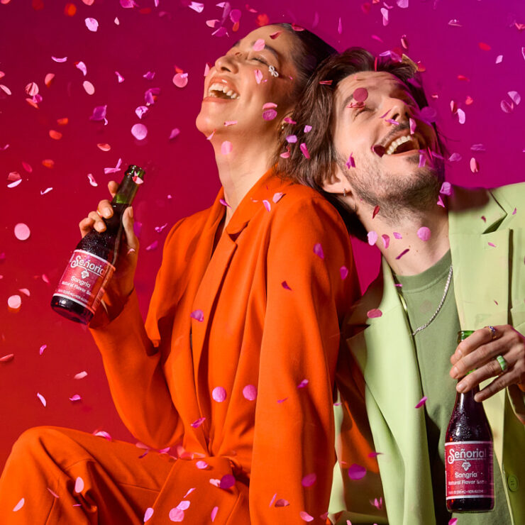 wo people celebrate joyfully under falling confetti, each holding a Sangría Señorial bottle against a vibrant red-to-pink background. Both smile and lean back in laughter, creating an energetic, festive atmosphere.
