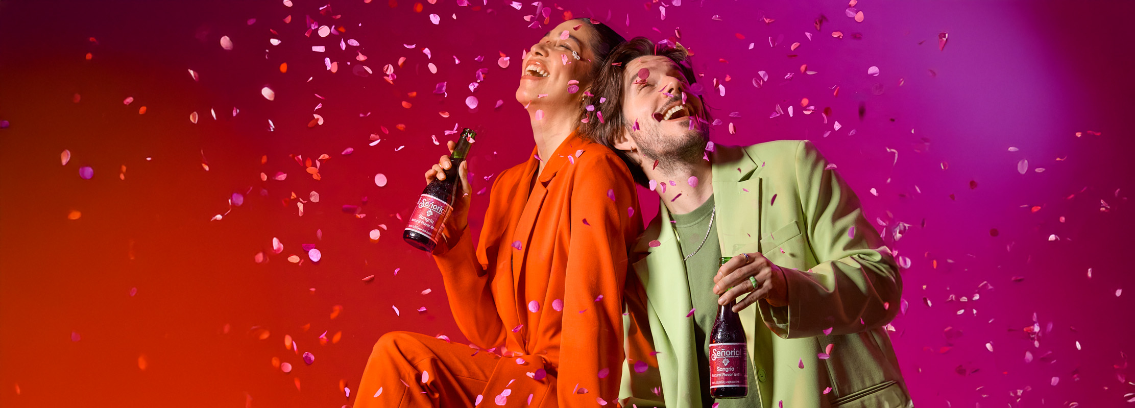 wo people celebrate joyfully under falling confetti, each holding a Sangría Señorial bottle against a vibrant red-to-pink background. Both smile and lean back in laughter, creating an energetic, festive atmosphere.