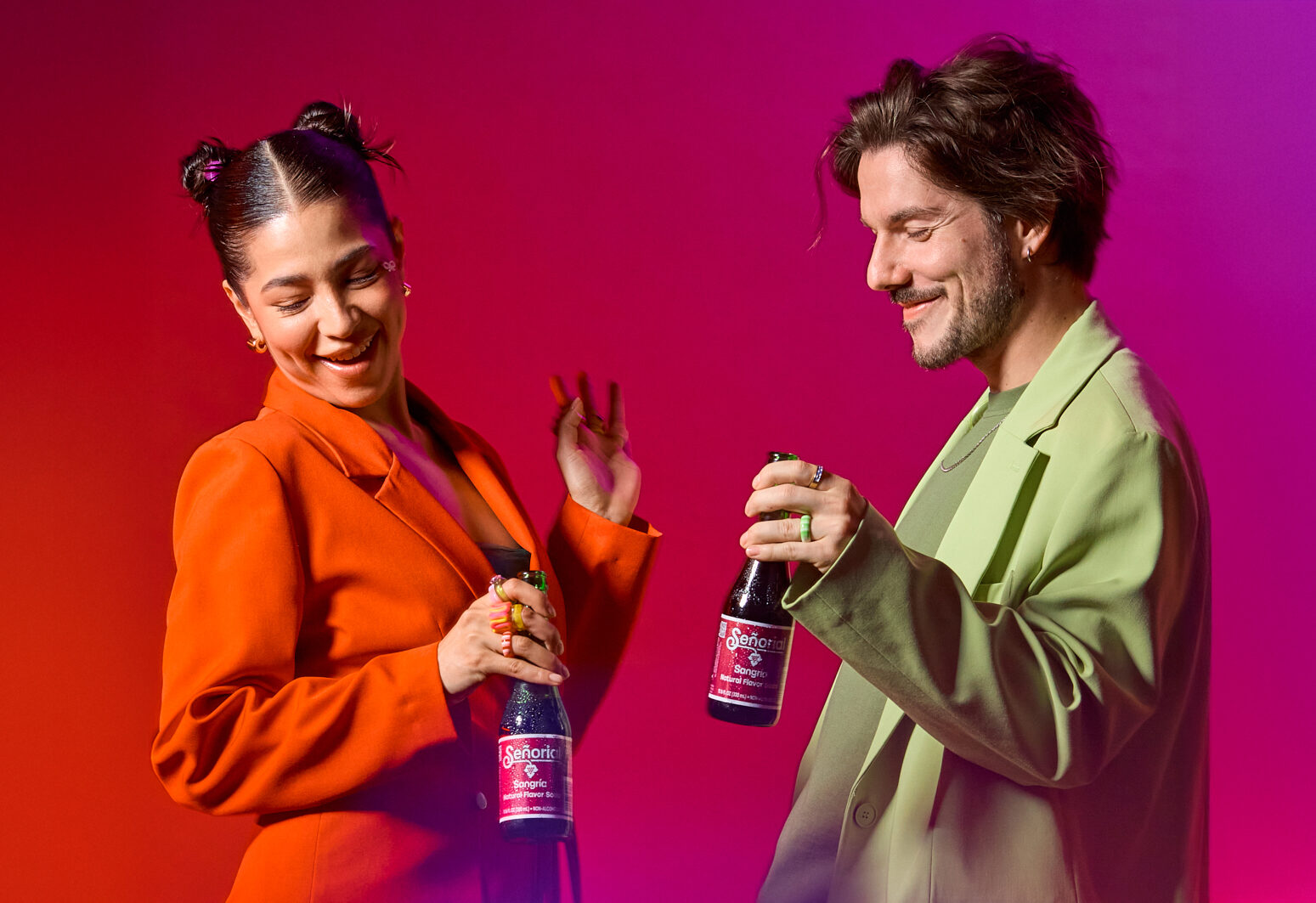 Two young adults smiling and dancing while holding bottles of Sangría Señorial, dressed in colorful blazers against a vibrant pink and red background.