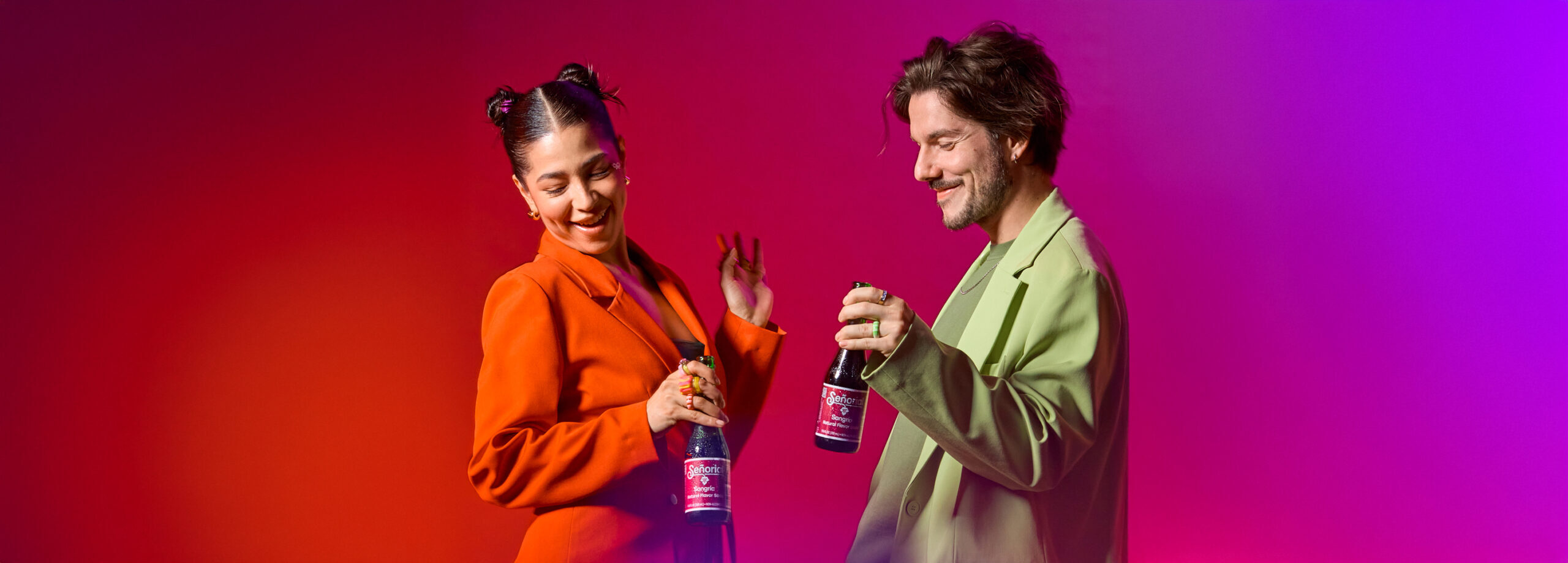 Two young adults smiling and dancing while holding bottles of Sangría Señorial, dressed in colorful blazers against a vibrant pink and red background.