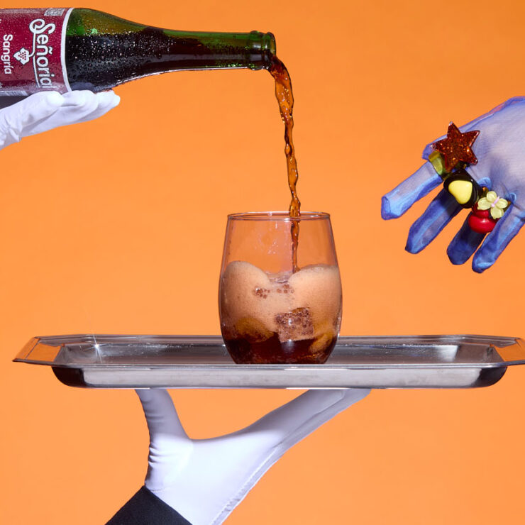“Hands pouring Sangría Señorial into a glass on a silver tray against a bold orange backdrop, creating a playful, elevated serving moment.