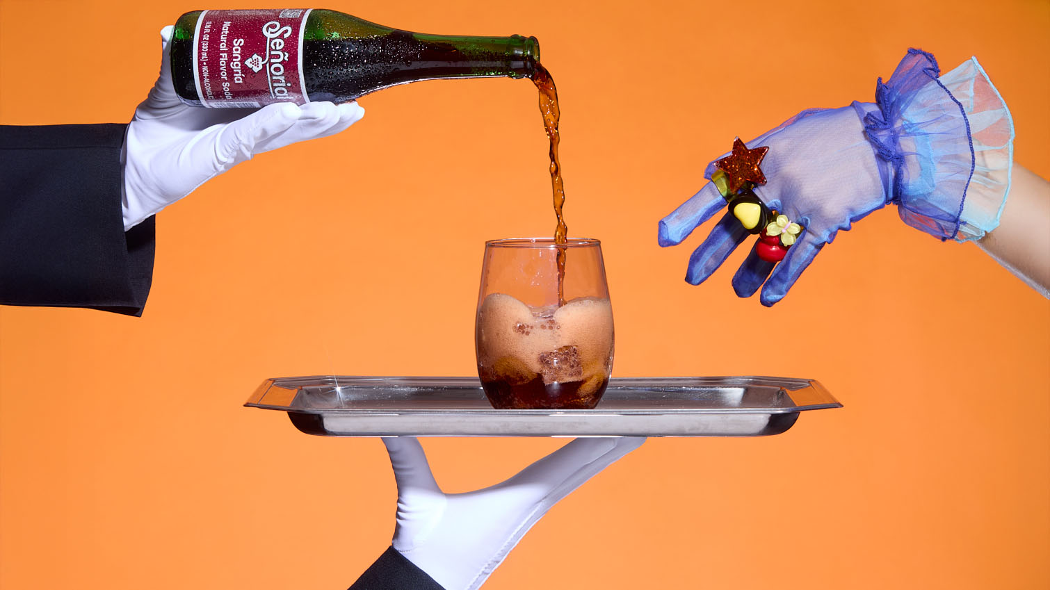 “Hands pouring Sangría Señorial into a glass on a silver tray against a bold orange backdrop, creating a playful, elevated serving moment.