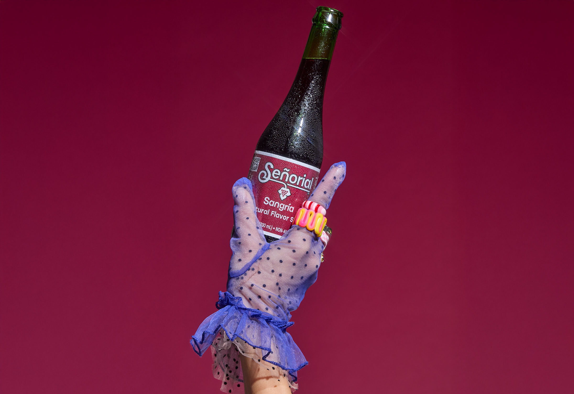 A stylish gloved hand holding a chilled Sangría Señorial bottle against a bold magenta backdrop.