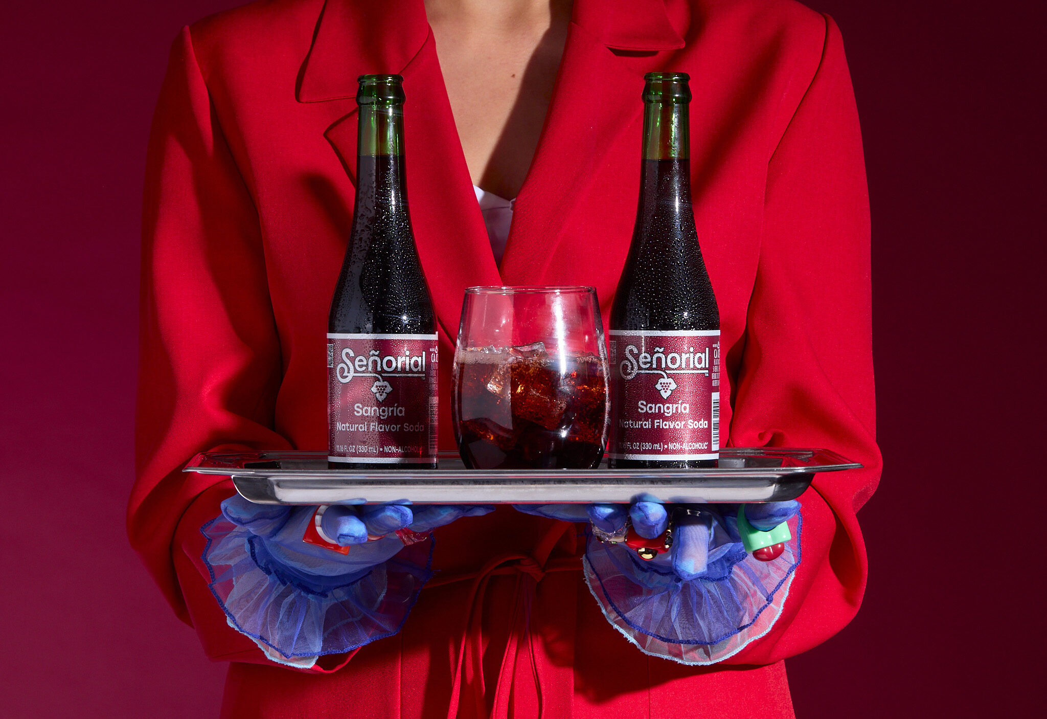 Two Sangría Señorial bottles and a glass on a serving tray, presented by someone in a bold red outfit with colorful gloves.