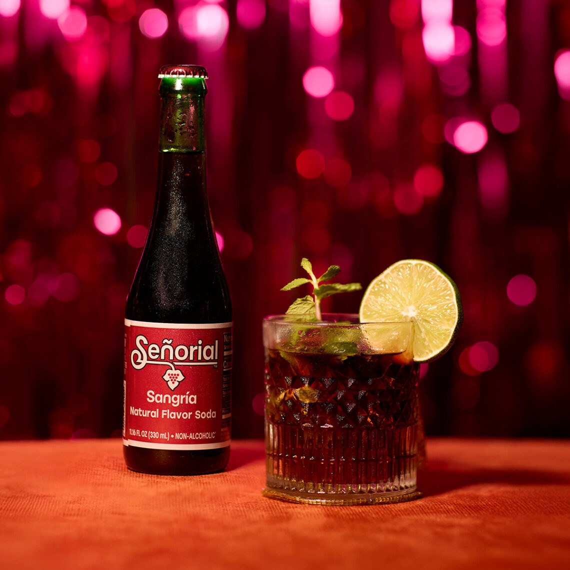 A bottle of Señorial Sangría soda beside a crafted sangría cocktail garnished with lime and mint, set against a festive red backdrop with sparkling bokeh lights.