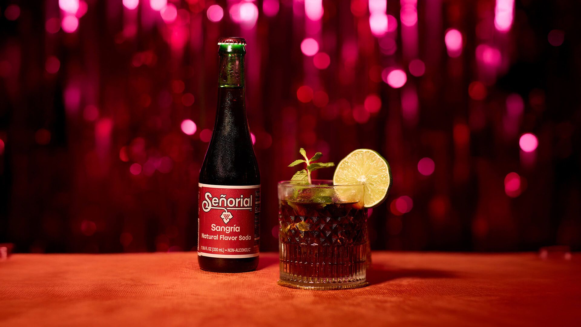 A bottle of Señorial Sangría soda beside a crafted sangría cocktail garnished with lime and mint, set against a festive red backdrop with sparkling bokeh lights.