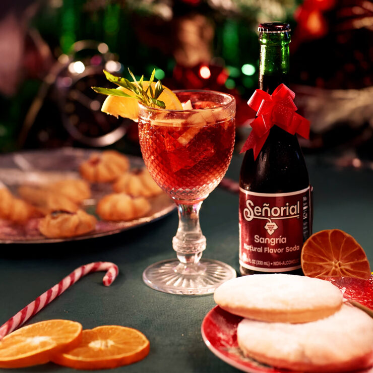 Festive holiday setup featuring a glass of sangría with fruit and herbs beside a Señorial Sangría soda bottle decorated with a red bow, surrounded by cookies, citrus slices, and Christmas décor.
