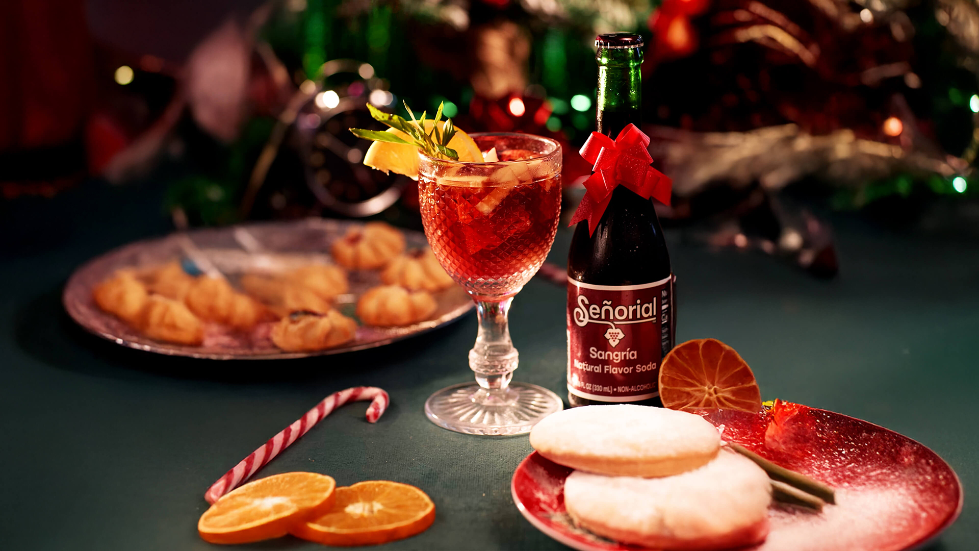 Festive holiday setup featuring a glass of sangría with fruit and herbs beside a Señorial Sangría soda bottle decorated with a red bow, surrounded by cookies, citrus slices, and Christmas décor.
