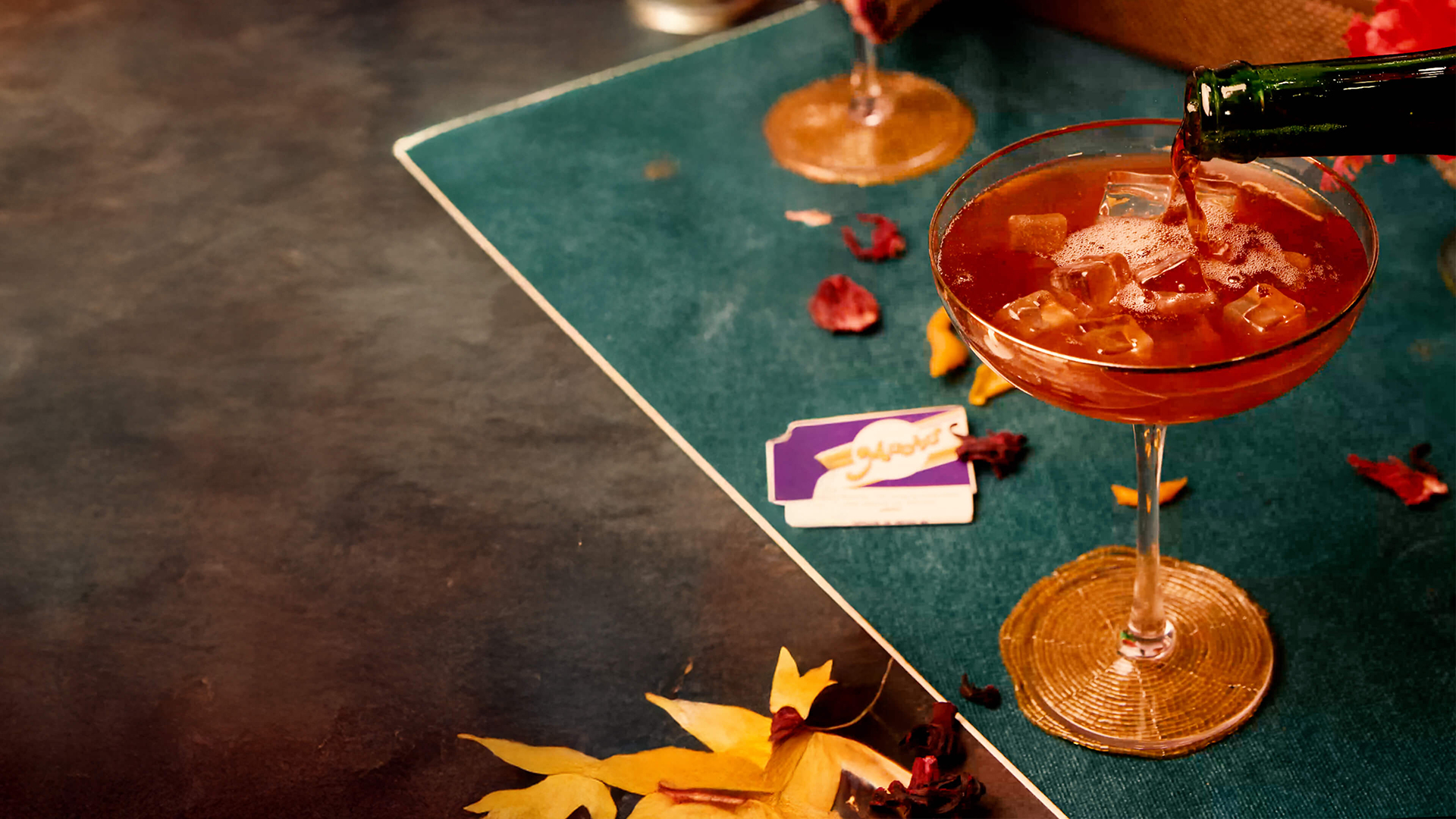 Pouring a sangría soda cocktail over ice in a coupe glass, surrounded by scattered dried petals on a teal table.