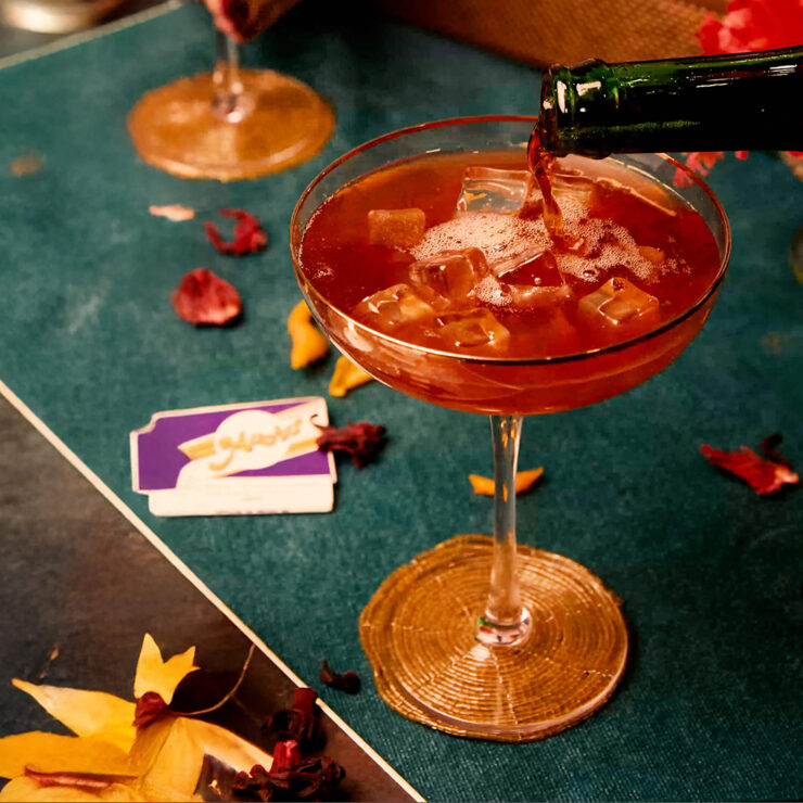 Pouring a sangría soda cocktail over ice in a coupe glass, surrounded by scattered dried petals on a teal table.