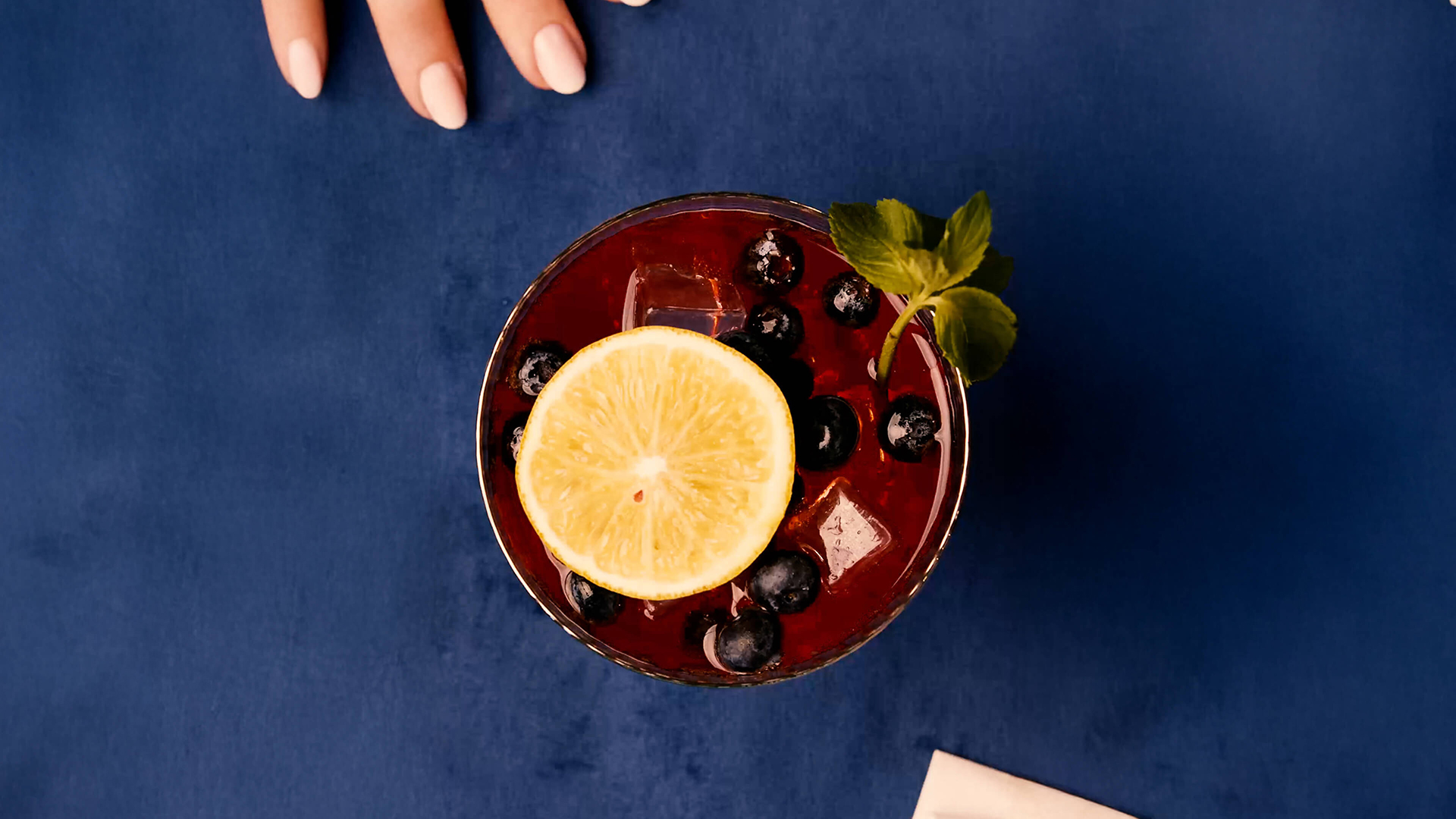 A refreshing sangría cocktail topped with blueberries, a lemon wheel, and fresh mint, styled on a bold blue background.