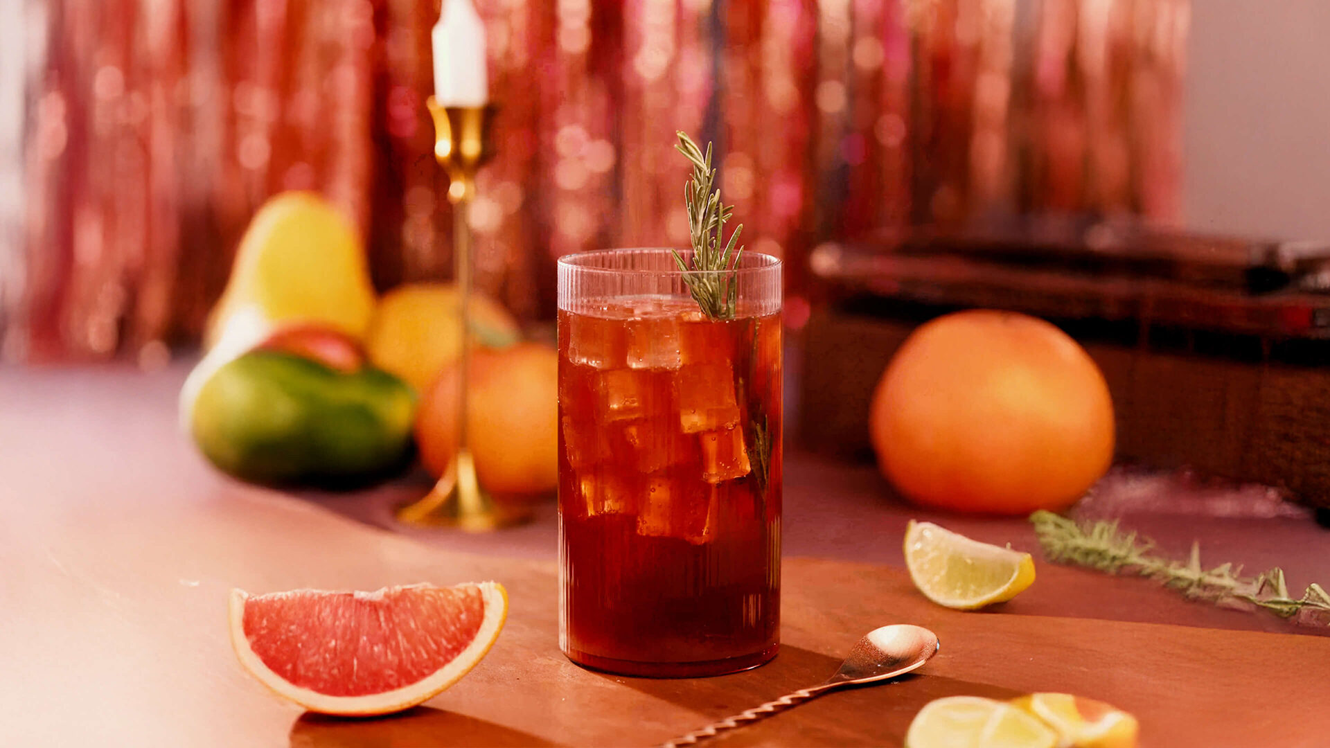 Tall glass of iced sangría soda with rosemary garnish, surrounded by citrus slices and fresh fruit on a warm, festive table.