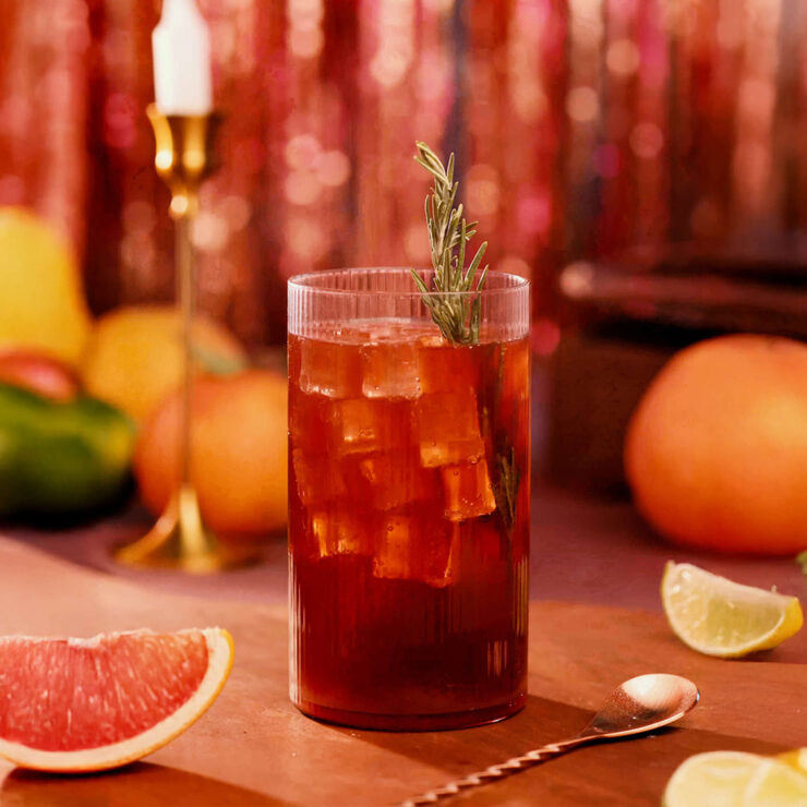 Tall glass of iced sangría soda with rosemary garnish, surrounded by citrus slices and fresh fruit on a warm, festive table.