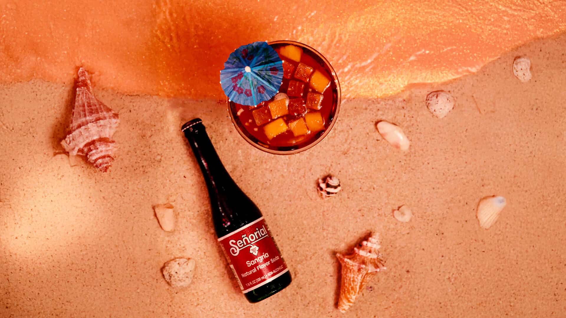 Sangria Senorial bottle and tropical mocktail on sand with fruit and seashells, evoking a refreshing, premium summer moment.
