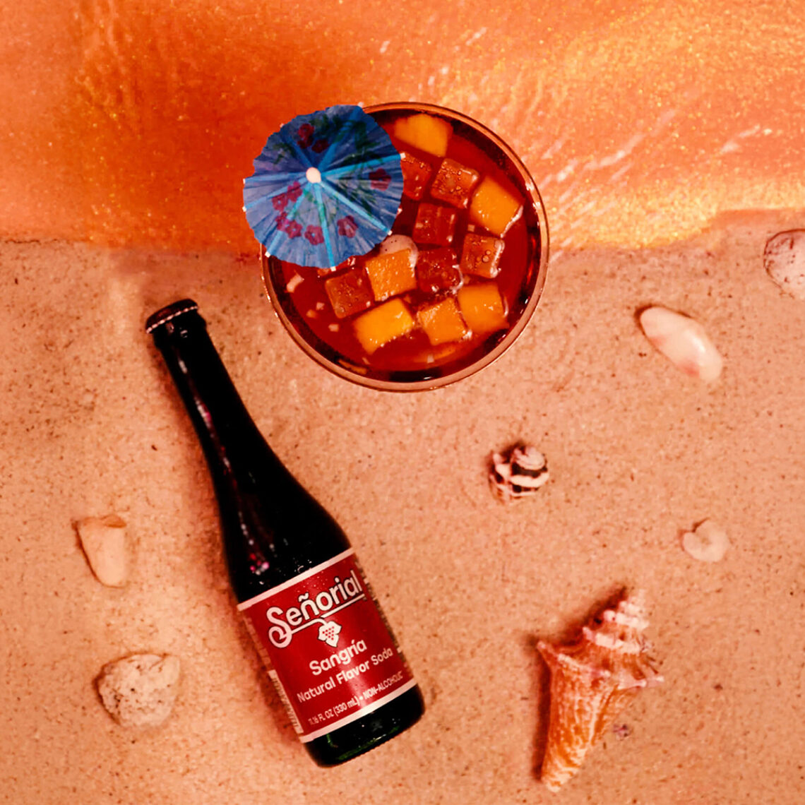 Señorial Sangría bottle on sandy beach beside a tropical sangría cocktail with fruit chunks and a blue umbrella, surrounded by seashells and shoreline foam.