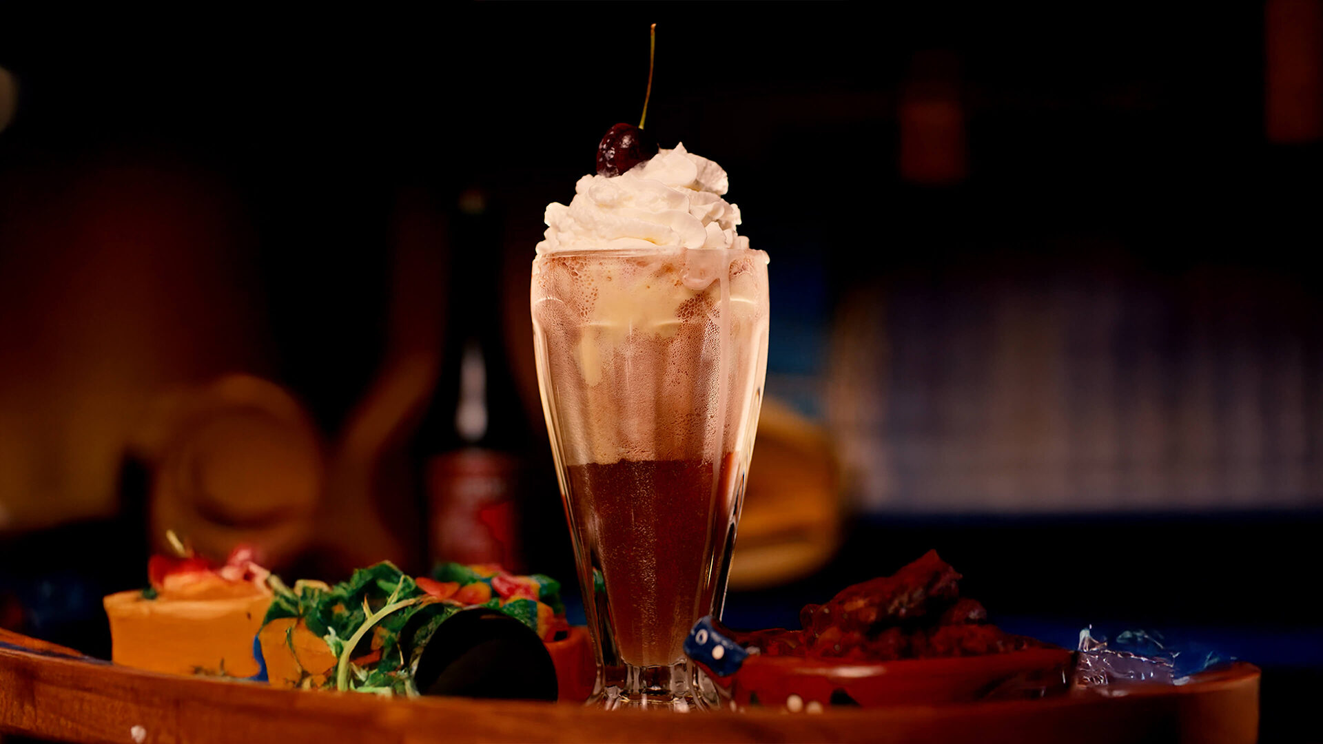 A tall ice-cream float made with sangría soda, topped with whipped cream and a cherry, surrounded by colorful garnishes and snacks on a warm, cozy table setting.