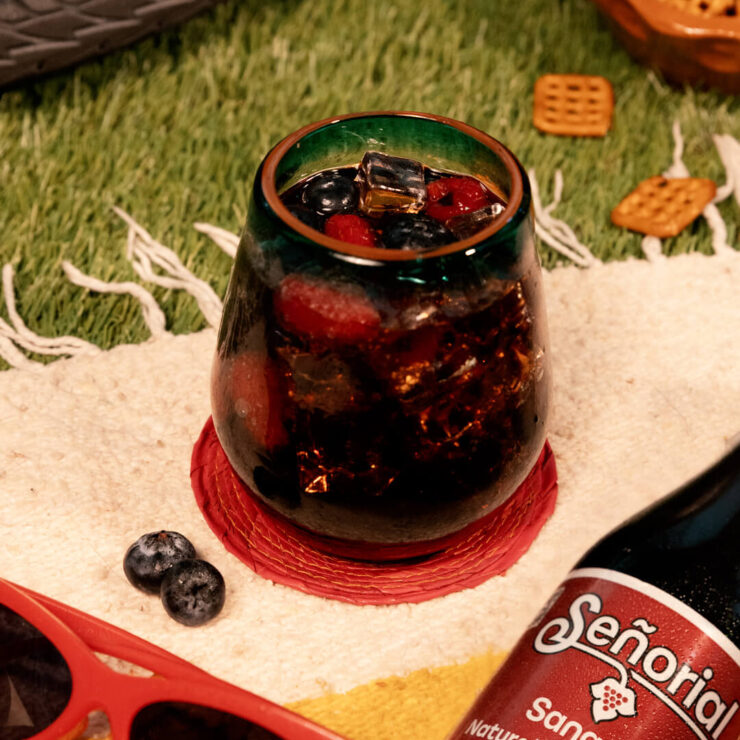 Glass of sangría soda with berries and ice on a picnic blanket, surrounded by sunglasses, blueberries, and a Señorío soda bottle.