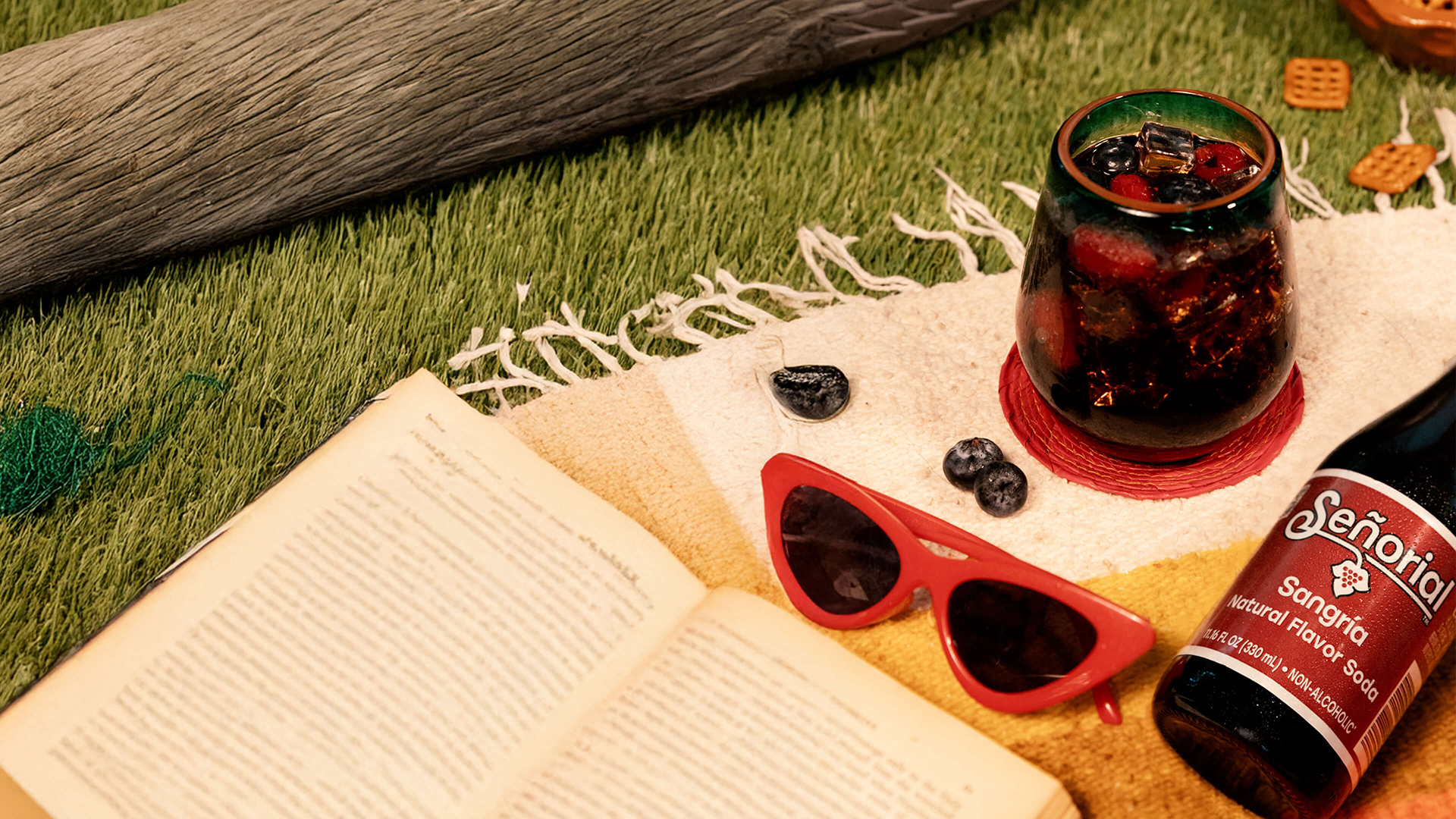 Glass of sangría soda with berries and ice on a picnic blanket, surrounded by sunglasses, blueberries, and a Señorío soda bottle.