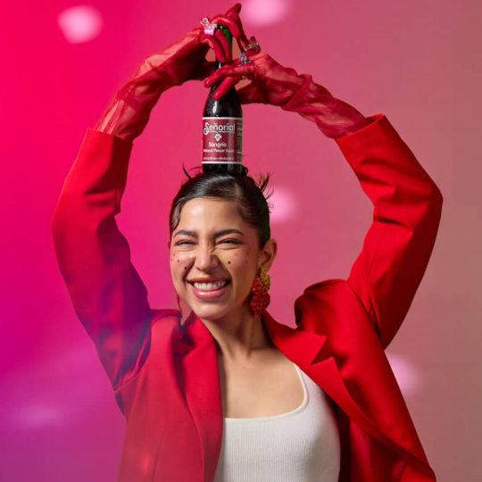Joyful lifestyle portrait featuring Sangria Senorial bottle held overhead, showcasing bold color, playful energy, and a modern non-alcoholic brand vibe.